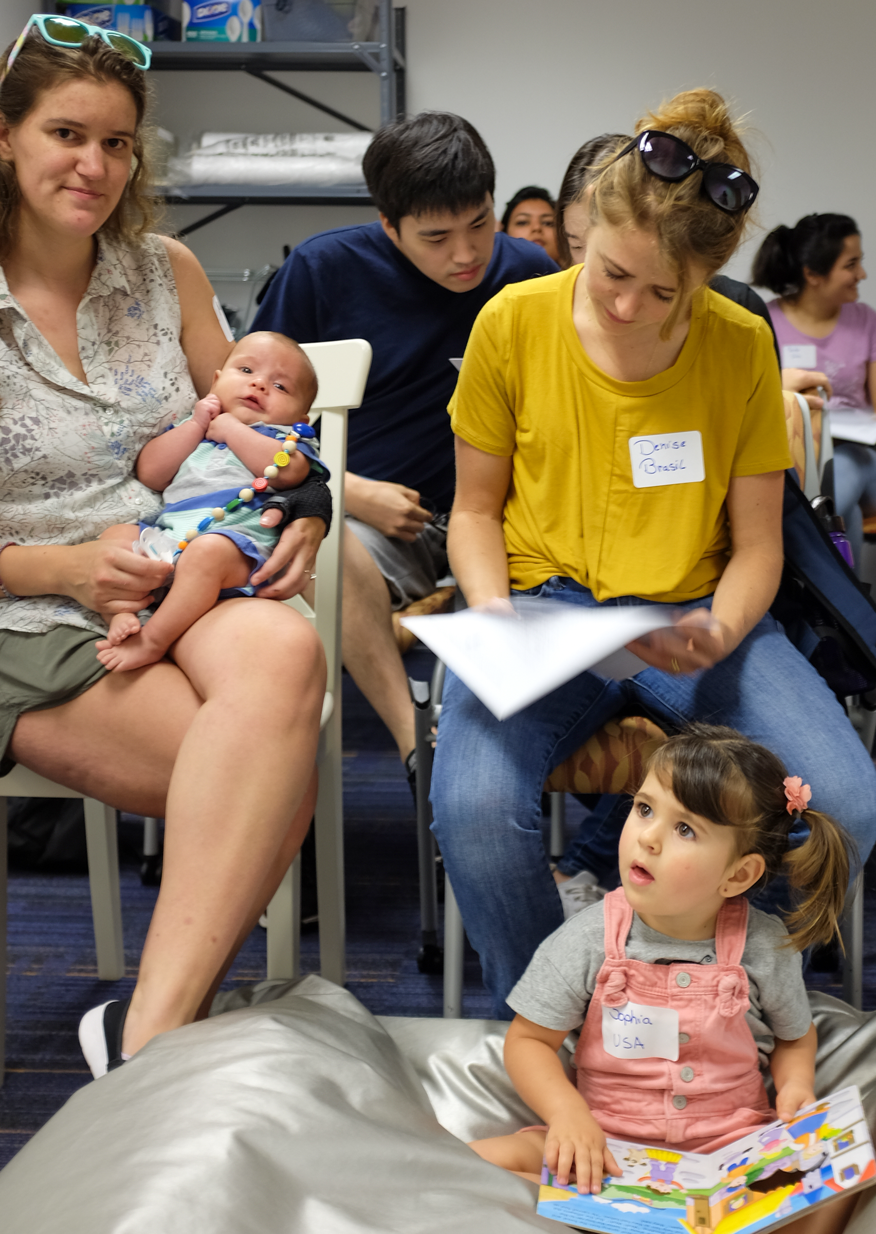 A woman holding a baby, another writing on a paper and a little girl reading a book sitting on a pillow on the floor.