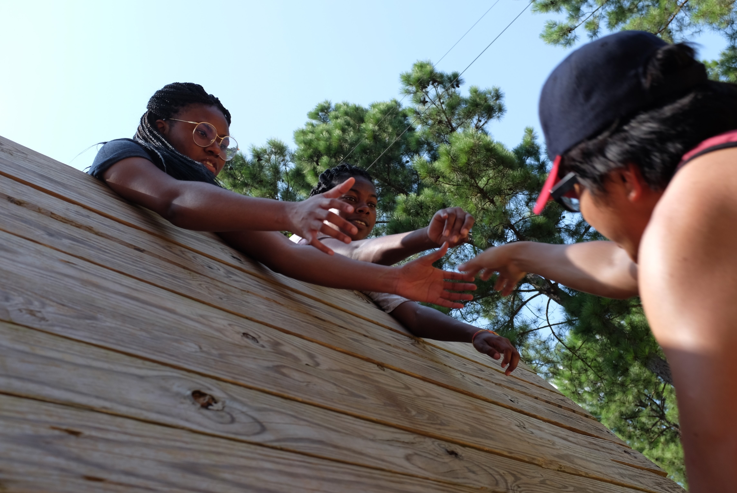 Two Black females helping a male go up a wall of an obstacle course.